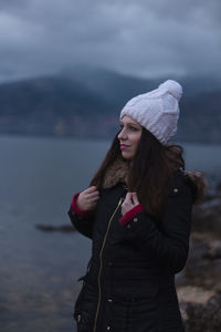 Thoughtful beautiful woman looking away while standing by lake during winter