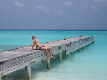 Full length of shirtless man sitting on pier over sea against sky