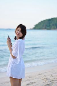Portrait of young woman with mobile phone standing on shore at beach against sky