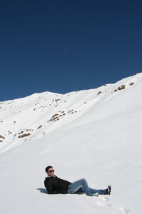 Full length of man lying down on snow covered landscape against clear blue sky