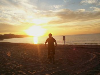 Man riding bicycle at beach against sky during sunset