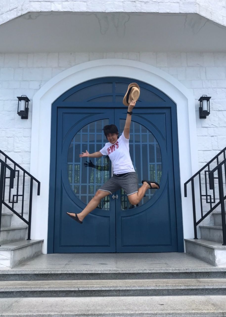 full length, one person, architecture, adult, young adult, casual clothing, built structure, blue, day, men, lifestyles, staircase, building exterior, window, leisure activity, person, white, door, entrance, city, arm, railing, mid-air, outdoors, women, wall, arms raised