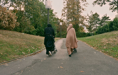 Rear view of women pushing baby strollers on road amidst trees