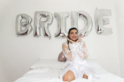 Woman sitting on bed against white wall with writing bride.