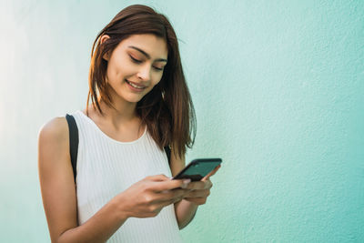 Young woman using phone while standing against wall