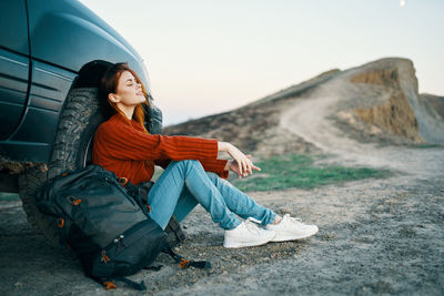 Young woman looking away while sitting on land against sky