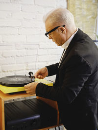 Side view of senior male in elegant jacket standing near table with retro record player and putting vinyl disc for listening to music