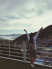 Full length of woman with arms raised standing by railing against sky