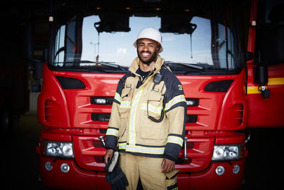 Portrait of smiling firefighter in uniform standing against fire engine at fire station