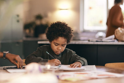 Boy with curly hair writing in book at home