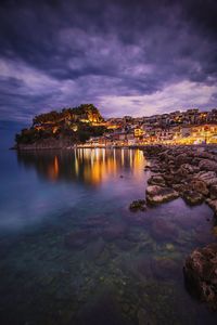 Illuminated city by sea against sky at sunset