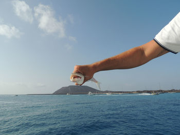 Cropped hand of man holding dead fish against sea and sky