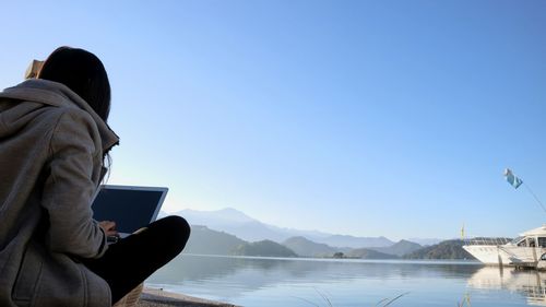 Woman using laptop by lake against clear blue sky