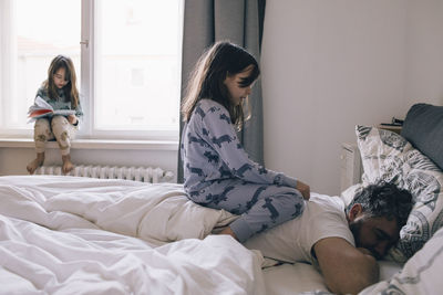 Girl sitting on father's back sleeping on bed while sister reading book at window