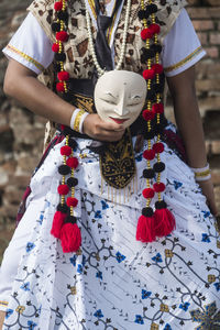 Midsection of woman in traditional clothing holding mask while standing against wall