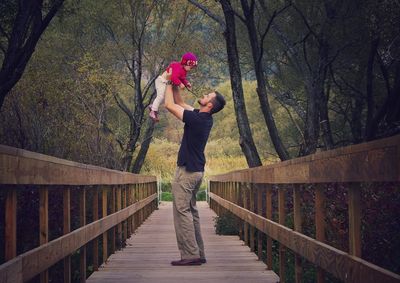 Full length of happy father carrying daughter amidst trees on wooden footbridge