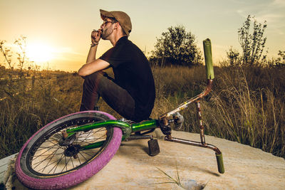 Man sitting with bicycle on field against sky during sunset