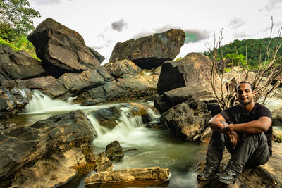 Portrait of man sitting on rock against sky