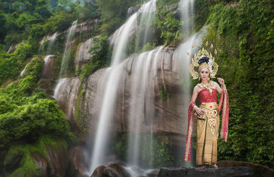 Portrait of beautiful young woman wearing crown and traditional clothing while standing against waterfall in forest