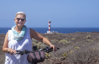Portrait of smiling senior woman with bicycle standing against lighthouse against sky