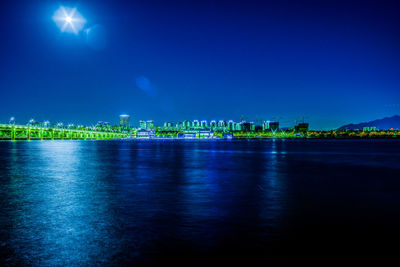 Illuminated city by sea against clear blue sky at night