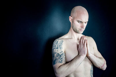 Shaved head shirtless man with hands clasped against black background