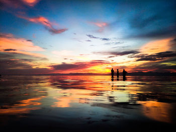 Silhouette people in sea against dramatic sky during sunset