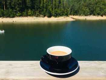 Cup of coffee placed on wooden table near a lake surrounded by green forest