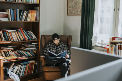 Young man reading book while sitting on armchair by bookshelf in library