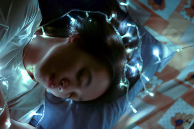 High angle view of woman with illuminated string lights lying on bed