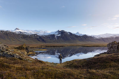 Back view of unrecognizable male tourist in casual clothes admiring wild nature while standing near peaceful lake reflecting snowy mountains in iceland