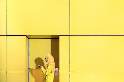 Side view of teenage girl using phone while standing by yellow wall