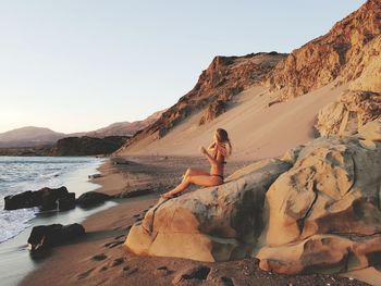 Side view of woman in bikini sitting on rock at shore