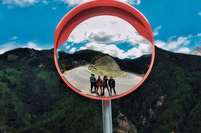Reflection of friends on traffic mirror by mountains against sky