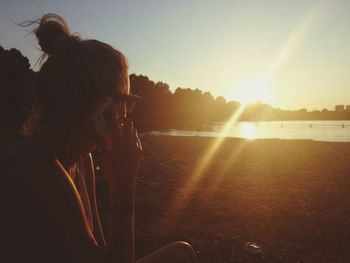 Woman answering smart phone while sitting on field by lake during sunset