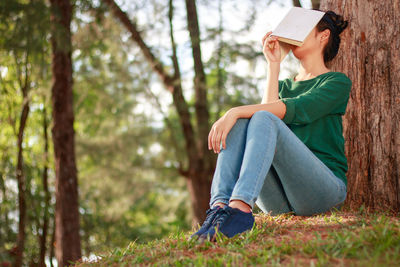 Full length of young woman covering face with book while leaning on tree at park
