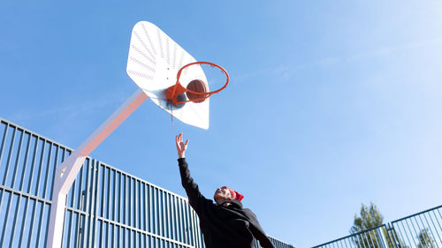 Active man throwing ball into basketball hoop with raised arm while playing on sports ground on sunny day during training