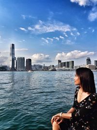 Mid adult woman sitting by river against sky in city