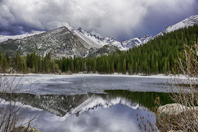 View of frozen lake with coniferous trees and snowcapped mountains against cloudy sky