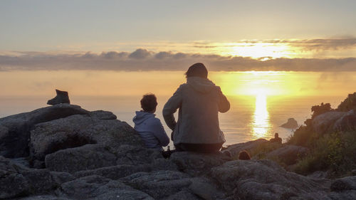 Rear view of people sitting on rock by sea against sky during sunset