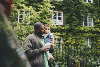 Affectionate father kissing son while standing against fence in city