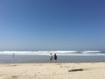 Rear view of couple standing at beach against clear blue sky