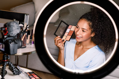 Smiling young woman applying make-up while being filmed in camera