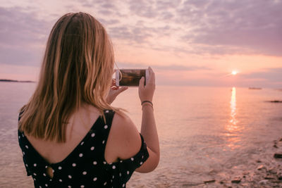 Back view of young woman taking a photo of sunset