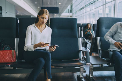 Young businesswoman text messaging through smart phone while sitting with passport in waiting room at airport
