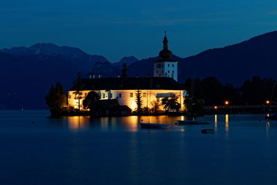 Illuminated building by lake against sky at dusk