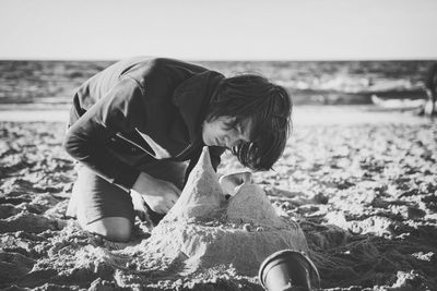 Teenage boy making sand castle at beach against sky