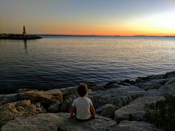 Rear view of boy sitting on rock by sea against clear sky
