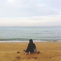 Rear view of young woman with cat sitting on sand at beach against sky