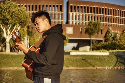 Side view of young man playing guitar while standing against building